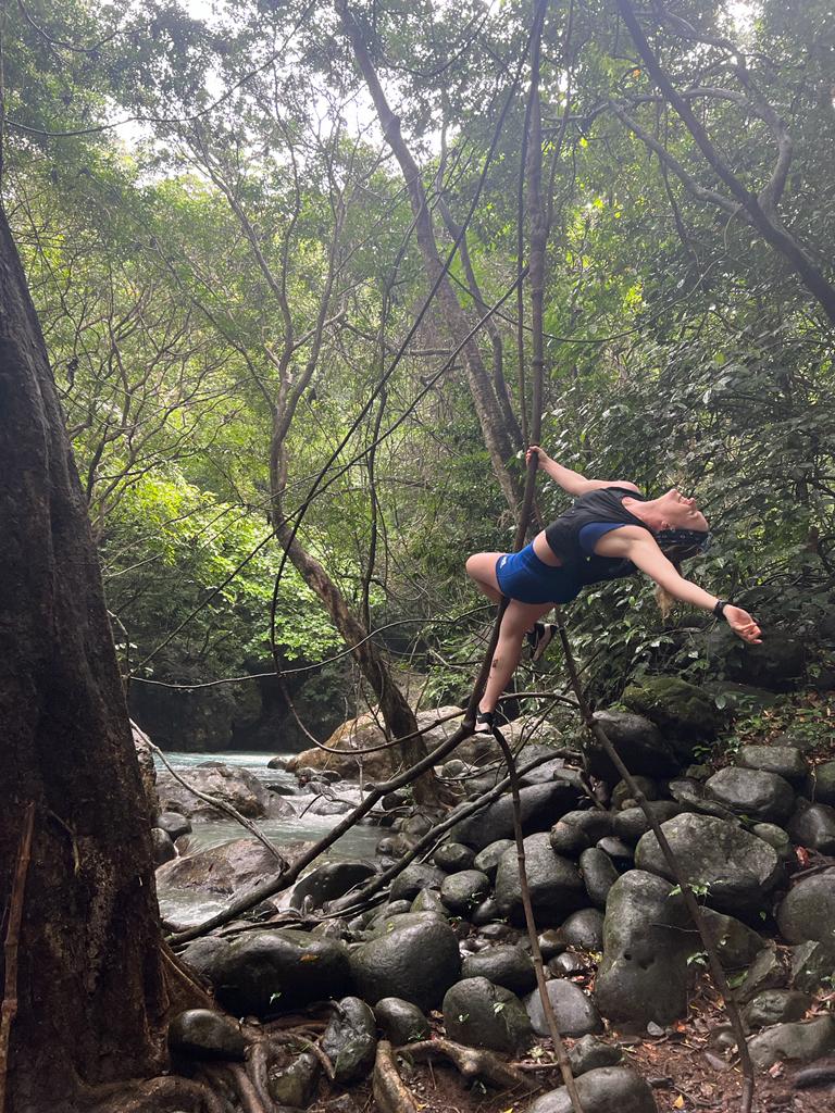 A woman hangs from thick vines above a rocky riverbed, legs hooked around one vine, torso arched backward and one arm reaching out. She wears a black crop top and blue shorts, with white-water rapids behind her and smooth dark boulders below